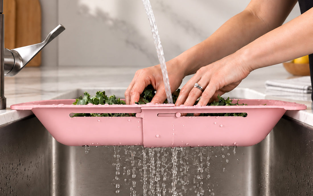 Pink Blue Ginkgo over-the-sink colander resting across a kitchen sink while hands rinse produce.