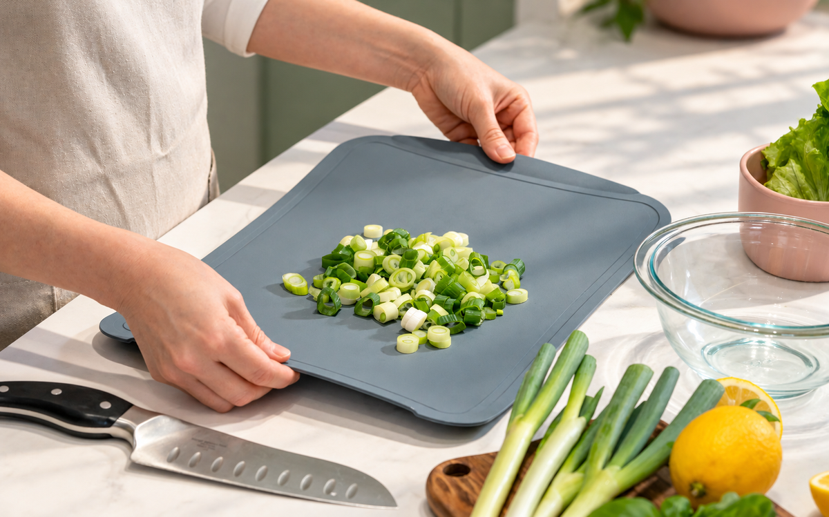 Person holding a flexible blue-gray TPU cutting board with chopped green onions beside a glass mixing bowl on a kitchen counter.