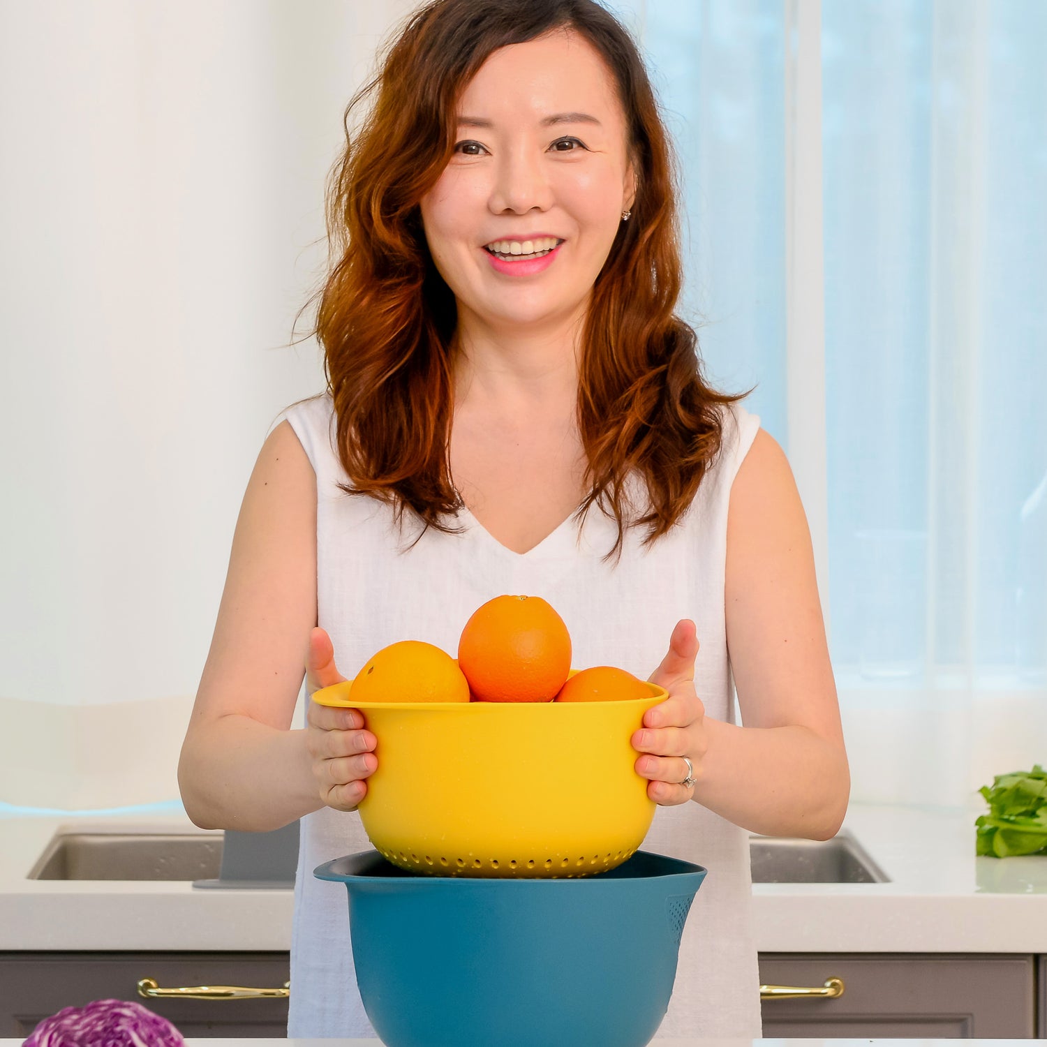 Woman holding a blue and yellow colander with oranges in a kitchen setting