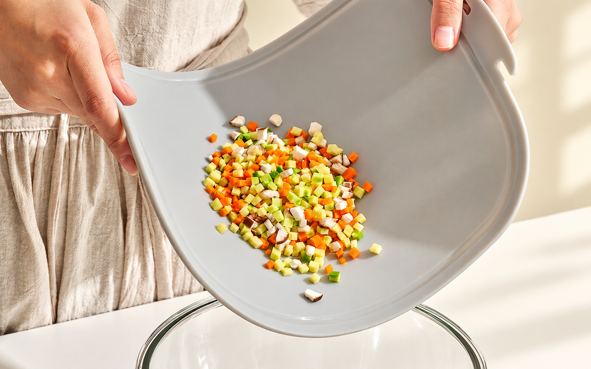 Hands bending a light gray Blue Ginkgo cutting board to guide diced vegetables into a glass mixing bowl.