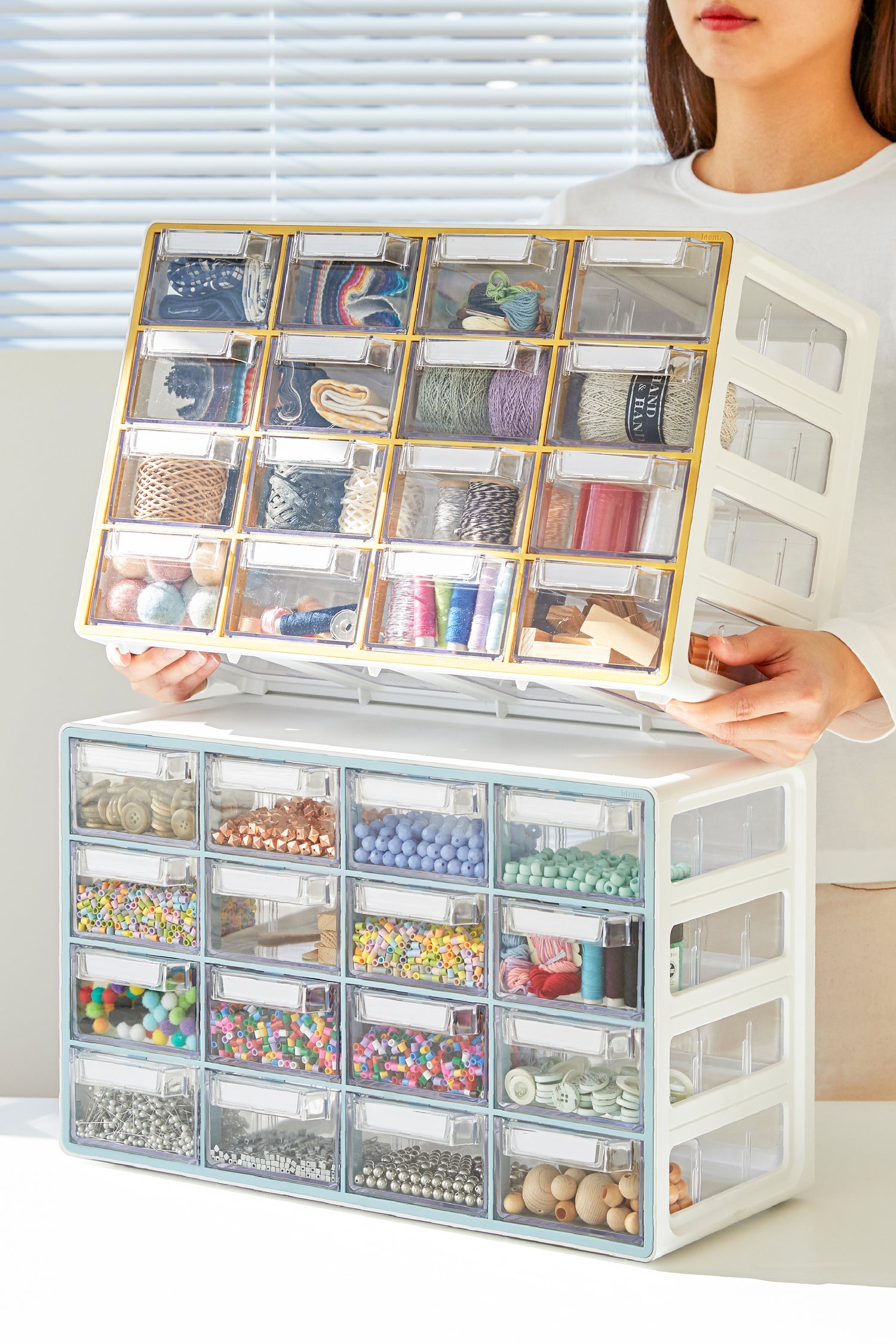 A lady places a 16 drawer storage cabinets on top of another. The cabinets feature a clear drawers used to store various items.