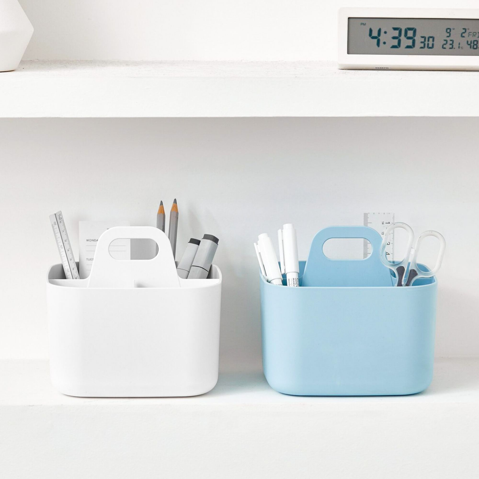 Two square caddies. One white and one blue, filled with office supplies on a light gray surface.